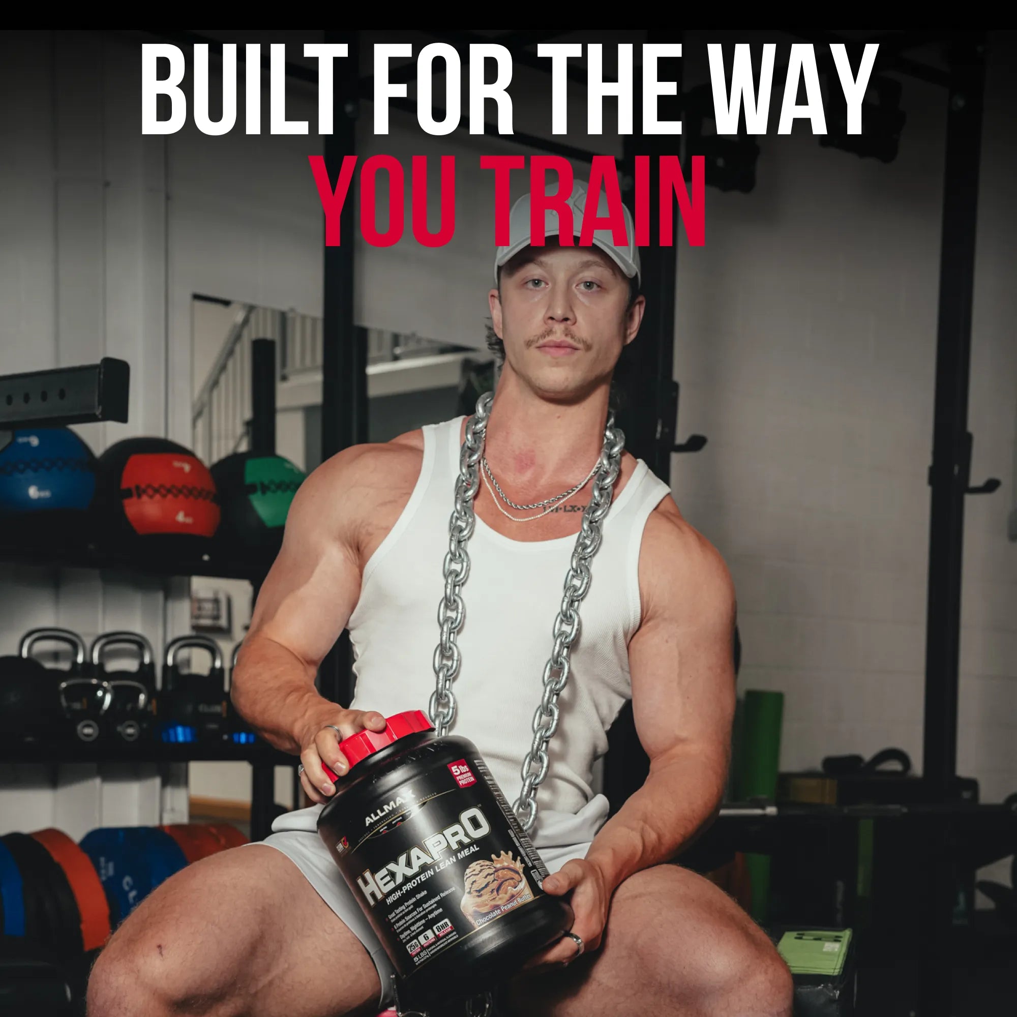 A muscular man in a gym setting wearing a white tank top and holding a tub of 100% Natural Hexapro protein powder. He has a silver chain draped around his neck.