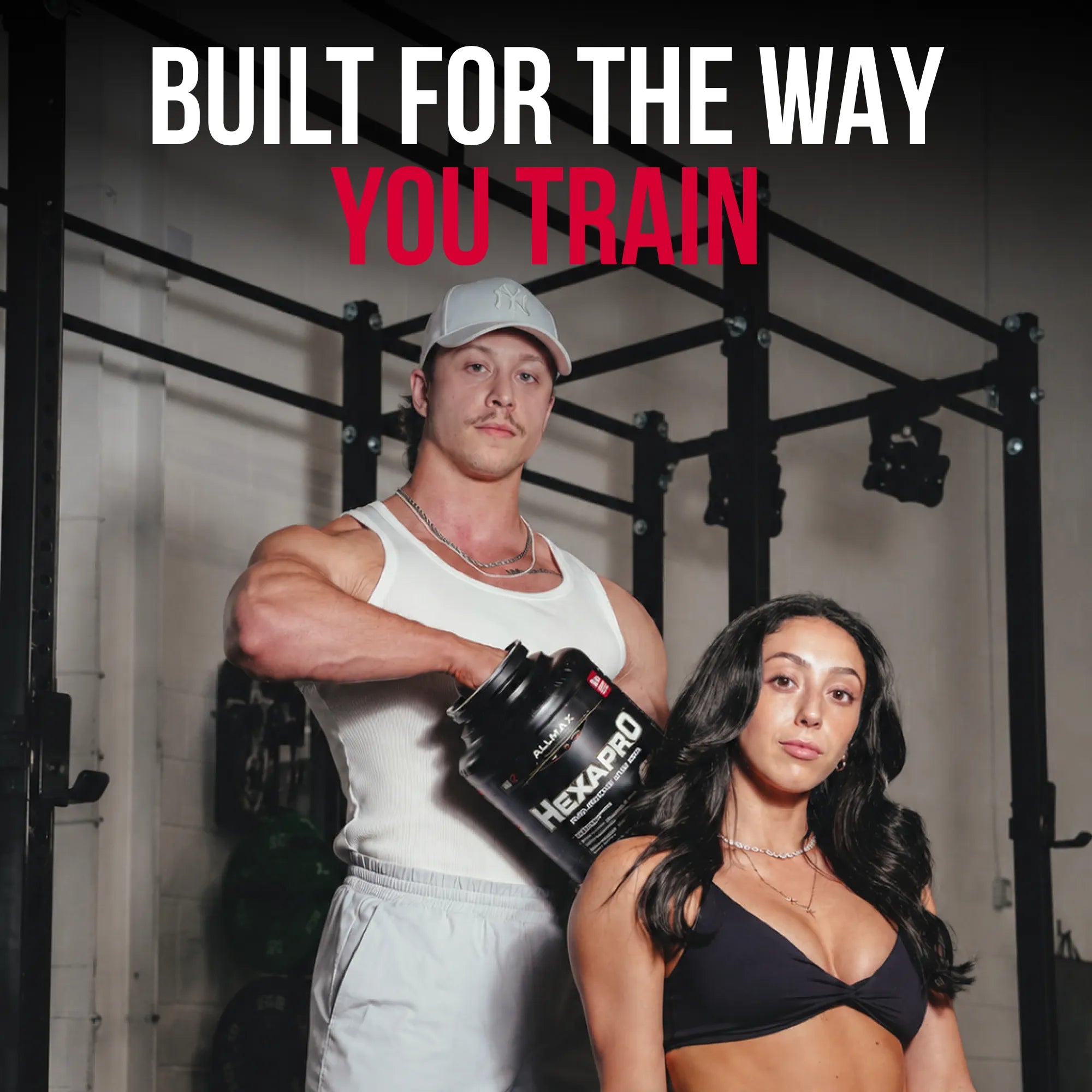 Two fitness models in a gym setting with a large tub of Hexapro pre-workout powder. The text overlay at the top reads 'BUILT FOR THE WAY YOU TRAIN' in white and red.